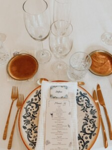 Primer plano de plato de cerámica decorado en azul con minuta de menú de boda, marcasitio personalizado y cubiertos dorados sobre mantelería blanca. Detalle de papelería de boda y bajo plato de cerámica artesanal